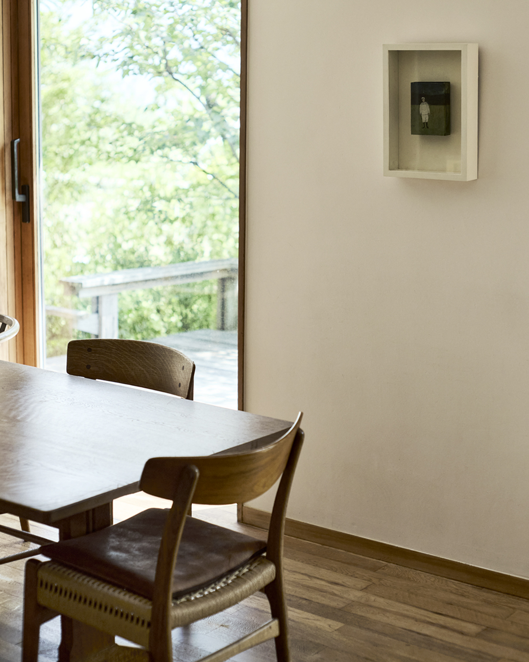 A corner of a table with some chairs with a picture frame on a white wall in a room within Ryuji Mitani's home.
