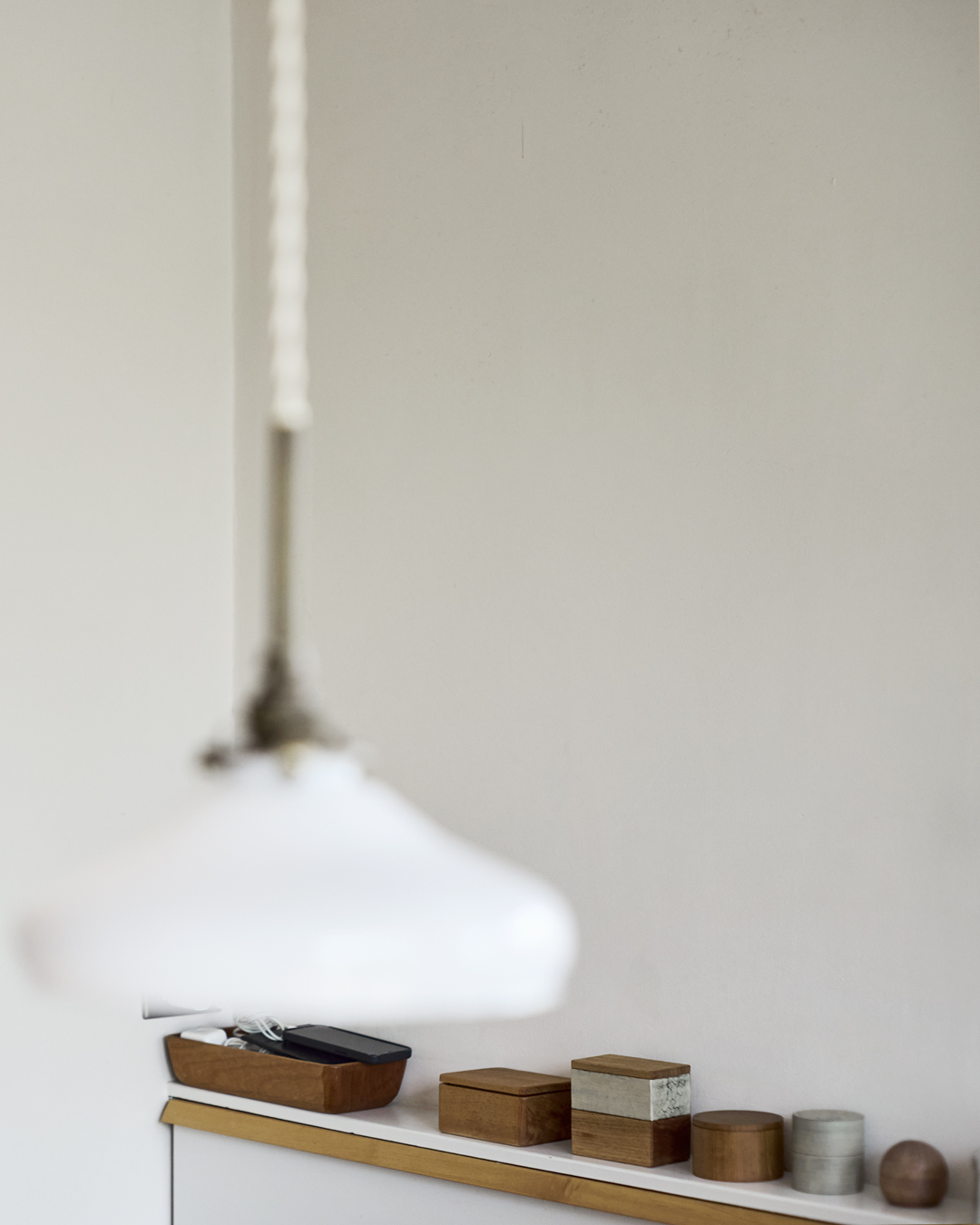 In a corner of a room, a pendant lamp shade is seen in a blurred foreground with wooden boxes and a tray lined on a shelf in the back in the camera's focus.