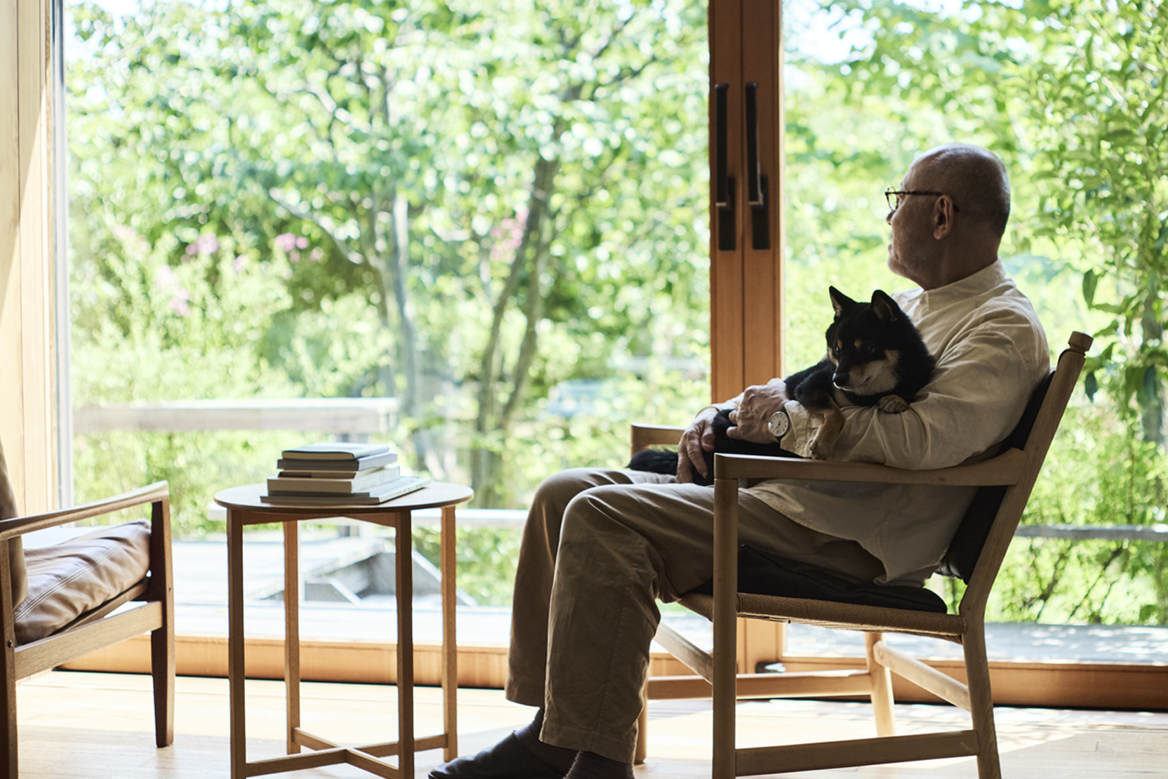 Ryuji Mitani sitting in a chair holding his dog and staring out a large window door.
