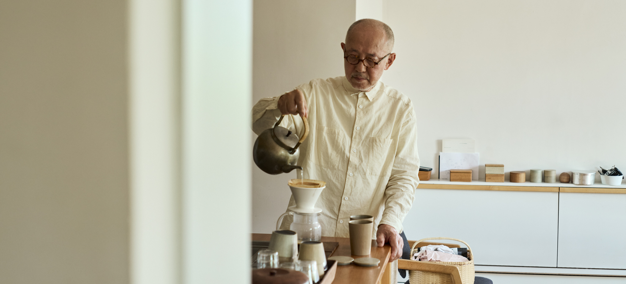 Ryuji Mitani pouring hot water into a coffee dripper with his coffee cup made by himself beside it. In the background, there's wooden containers lined all along the shelf.