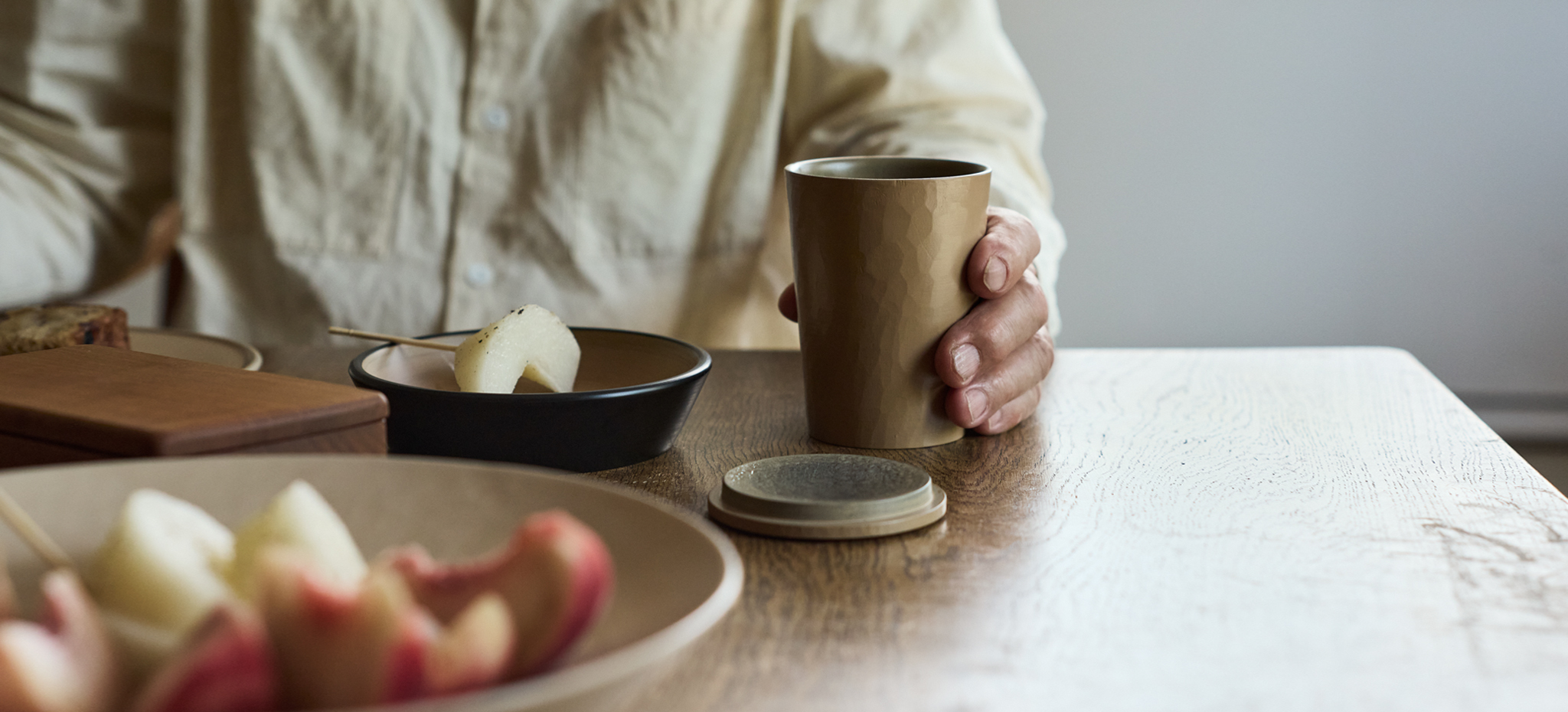 A close up of Ryuji Mitani 's hand holding a open wooden coffee cup with the lid laying on the side made by himself. Surrounding him are other wooden bowls and box,