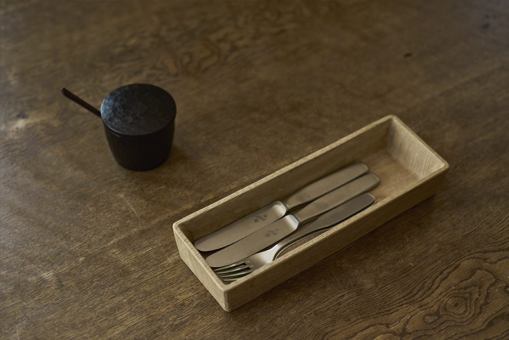 A wooden spice container with spoon and a wooden cutlery holder made by Ryuji Mitani on a wooden table top. The holder is filled with silver utensils.