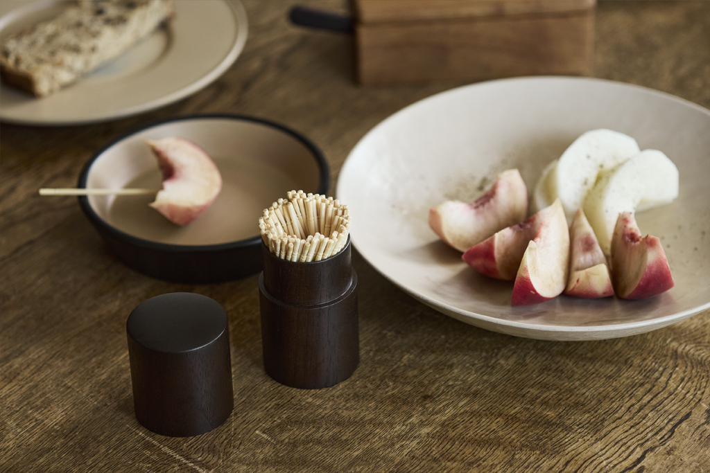 Wooden table top filled with wooden plates and food along side an open wooden toothpick container by Ryuji Mitani