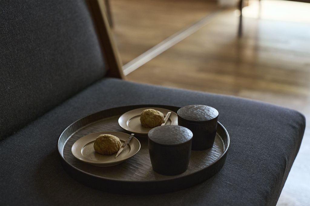 A wooden tray filled with wooden plates of pasteries and wooden lidded tea cups all sitting on a gray sofa