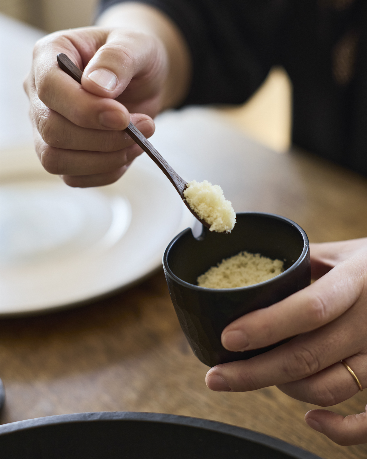 A person scooping a condiment with a wooden spoon out of a black wooden cup made by Ryuji Mitani