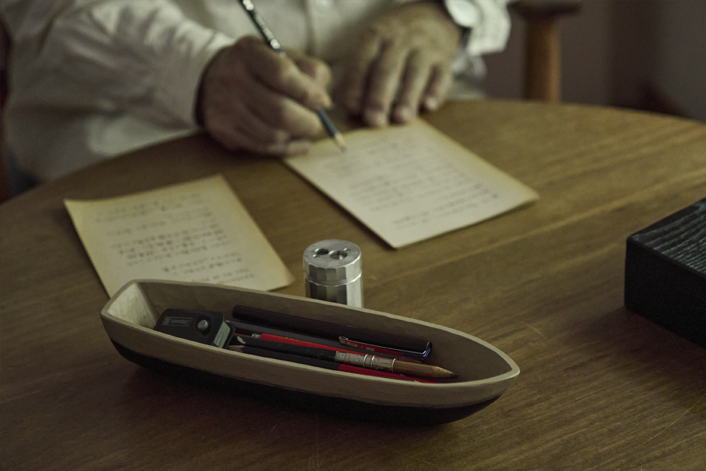 A tabletop showing a wooden boat stationary holder by Ryuji Mitani and displaying Mitani writing on paper in the back