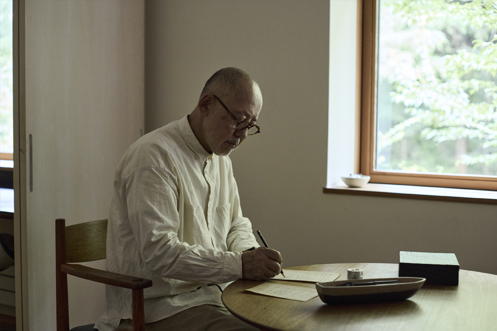 Ryuji Mitani sitting at a desk writing a letter with a stationery boat beside him